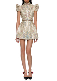 A model wearing a short, high-neck mini dress with puffed cap sleeves and an intricate floral brocade pattern in beige, gold, and pastel colors. The dress features a quilted diamond pattern and a thin beaded waistband.